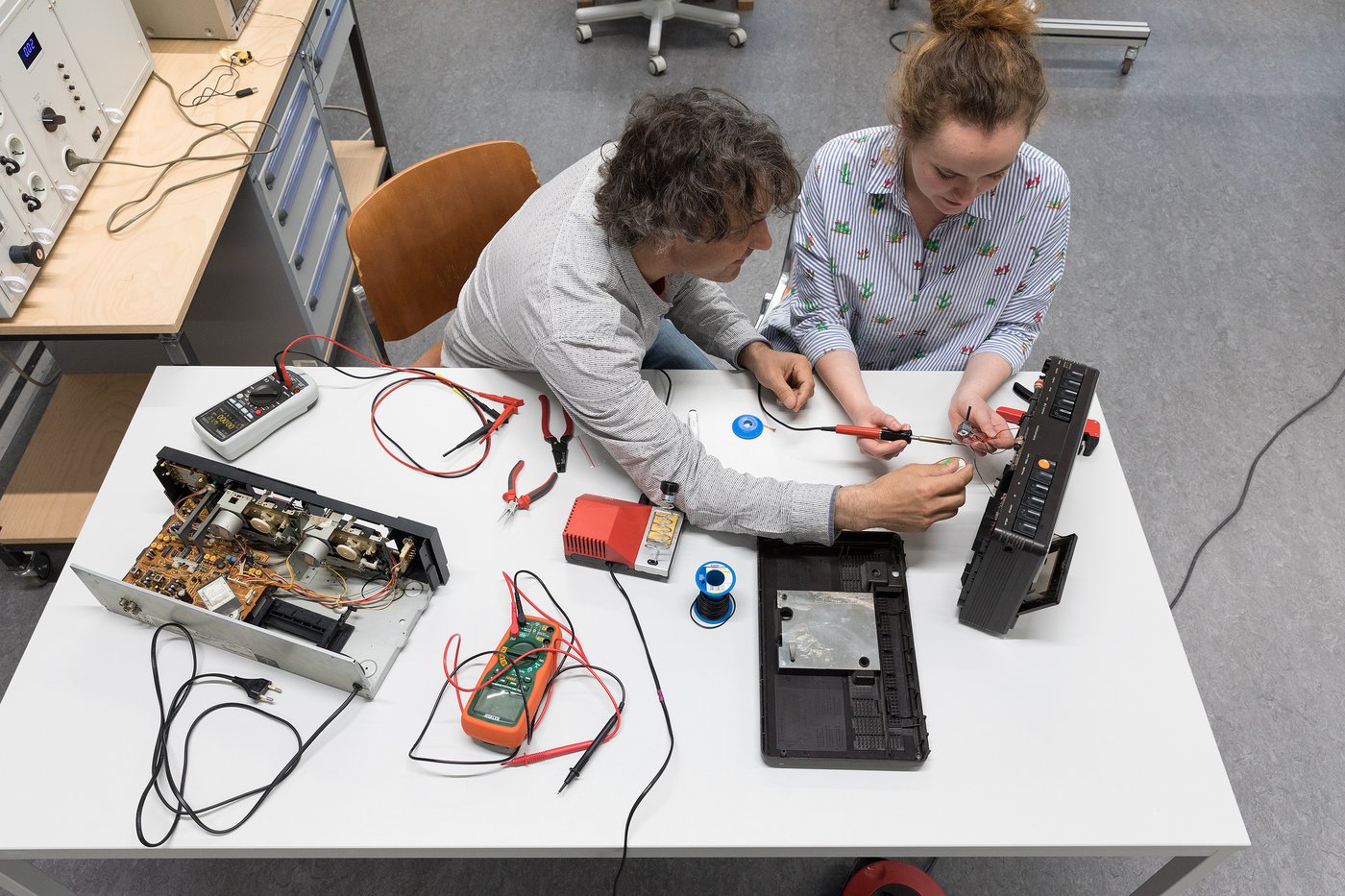 Im Bildvordergrund sieht man in Vogelperspektive einen hellen Arbeitstisch, auf dem ein auseinandergebautes Audio-Abspielgerät und Messgeräte liegen. Ein Lehrbeauftragter und eine Studentin sitzen nebeneinander an diesem Tisch und arbeiten gemeinsam an dem Gerät.