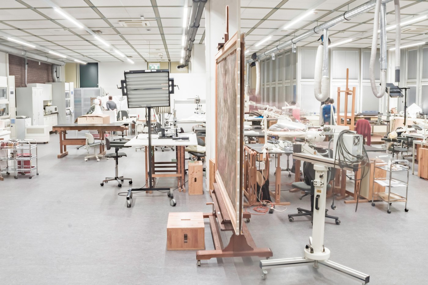 Blick in einen großen, hellen Atelierraum mit Fensterfront rechts. Es gibt ein paar Arbeitstische, Staffeleien und Mikroskope im Raum, im Hintergrund sind einige Studierende zu sehen.