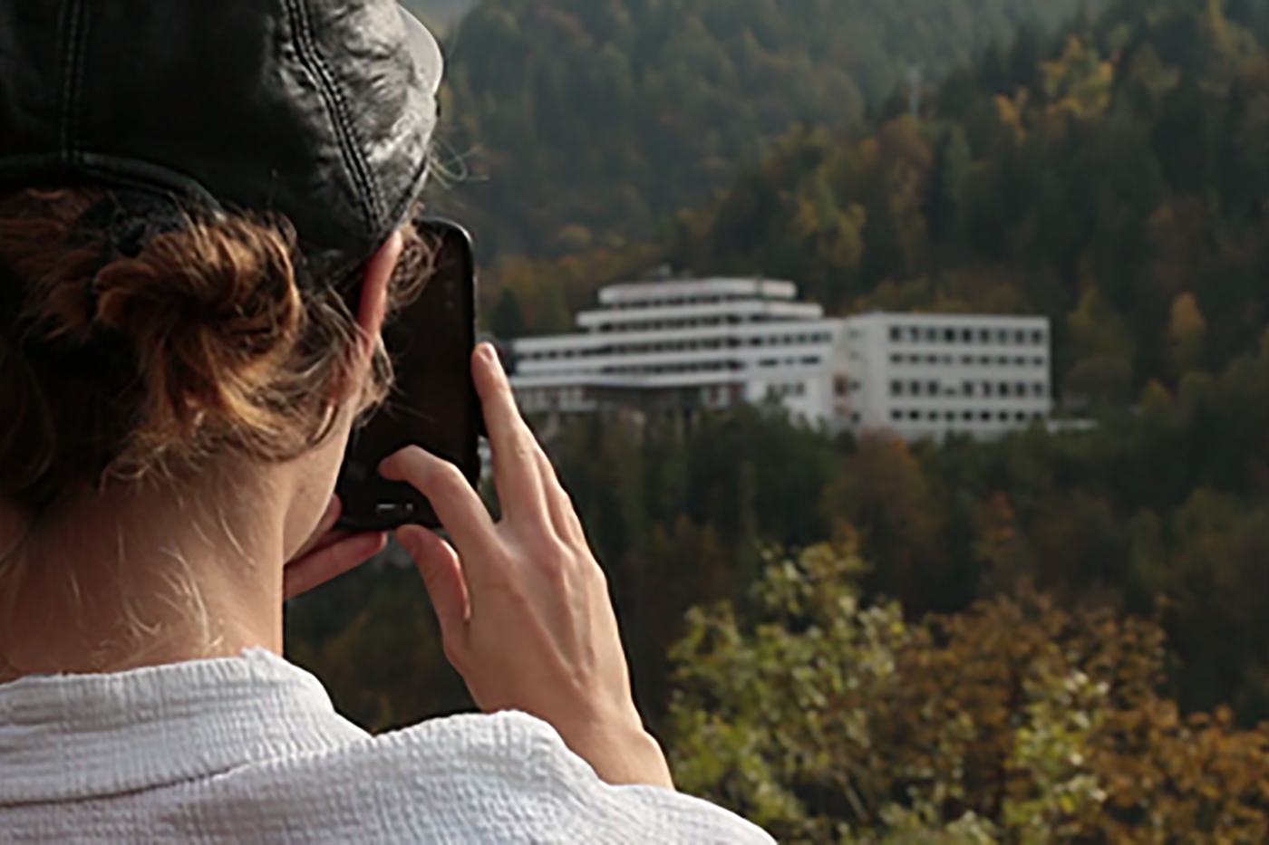 Videostill. Vordergrund links sieht man den Hinterkopf einer Person, die mit einem schwarzen Telefon ein Foto macht des Hintergrunds macht, wo in einem hügeligen Wald ein großes weißes Haus mit vielen Fenstern zu erkennen ist.