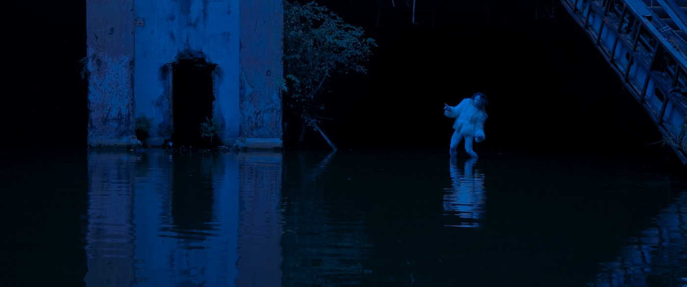 Ein weiß geschminkter und gekleidedeter Mann in einer Geste des Butoh-Tanzes steht in einem See innerhalb eines verfallenen Gebäudes.