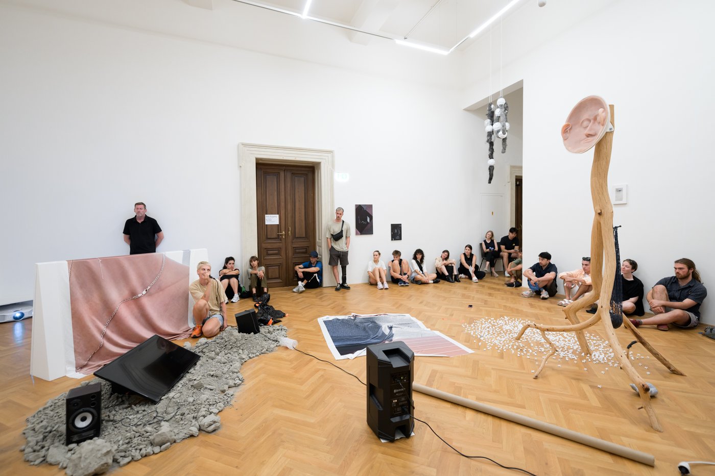 mehrere Personen in einem großen Raum mit Kunstwerken am Boden und an den Wänden, in der Mitte vom Raum findet eine Performance statt, Person sitzt am Boden mit Mikrofon in der Hand