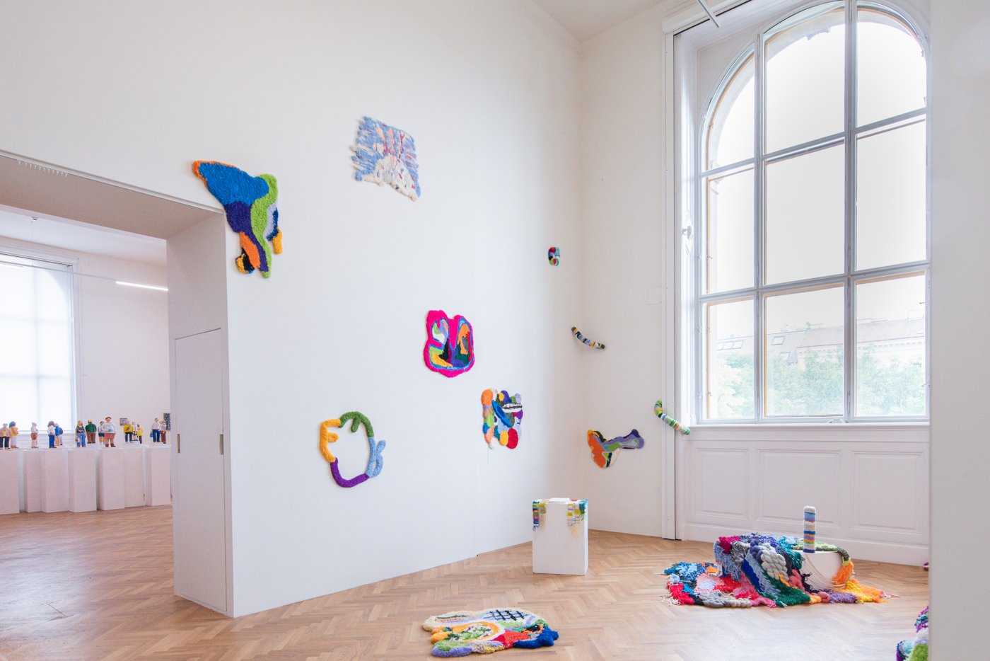 several colorful carpet-like fabric objects fixed on the floor and on the wall, on the right a large arched window, on the left the room opens up