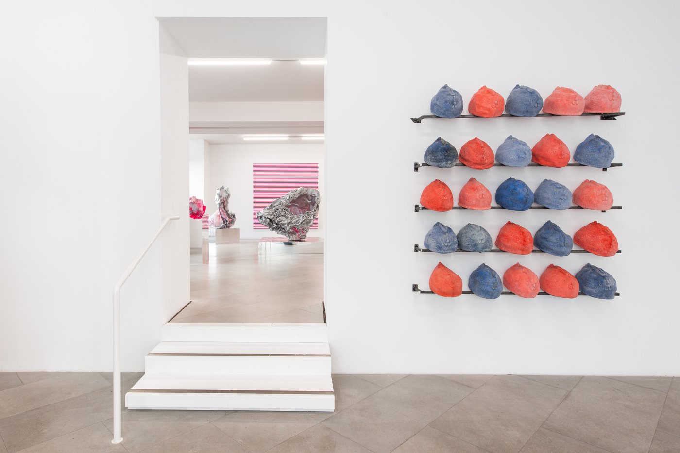 a room with two white steps and a white railing, on the right red and blue round objects with chicken faces lined up on black ledges, on the left view into the next room with silver-pink sculptures