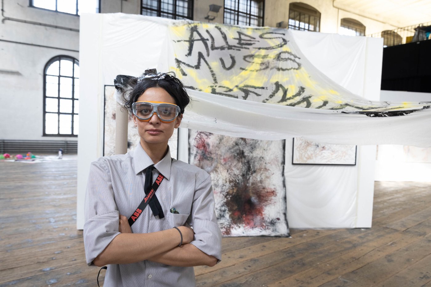 Frau blickt mit Sicherheitsbrille auf in die Kamera, hinter ihr ein Kunstwerk mit einem Banner "alles muss raus"