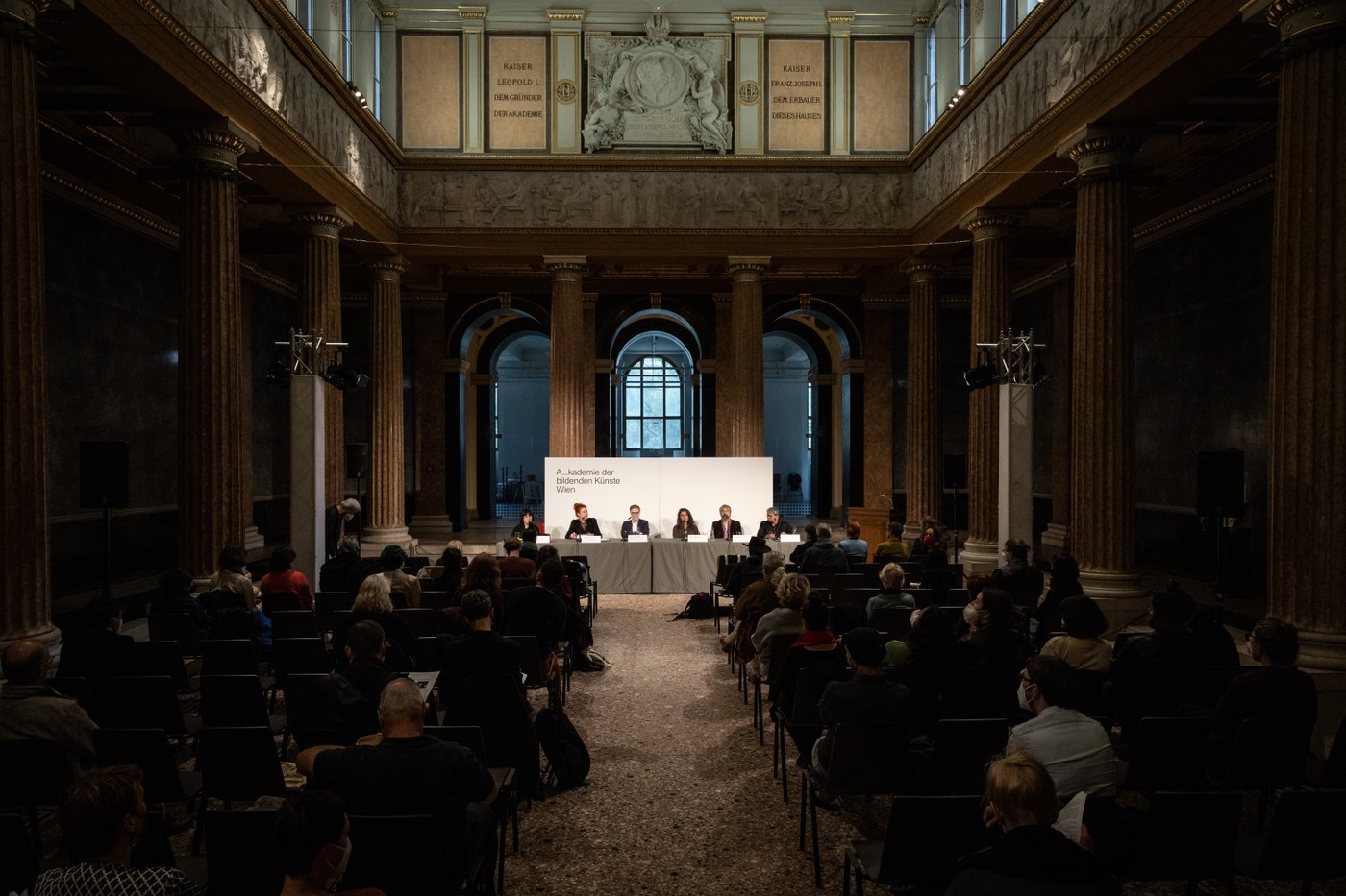 An einem langen Tisch in einem von Säulen eingefassten historischen Raum sitzen 5 Menschen, davor ein Publikum im Halbdunkeln.