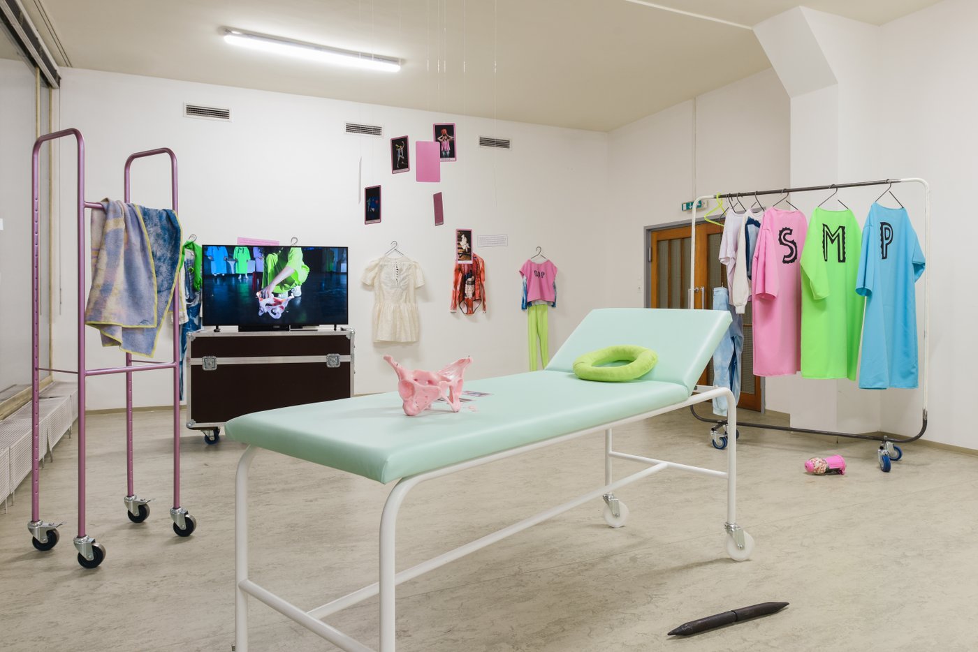 Ein Raum mit einer Krankenliege, einem gefüllten Kleiderständer und einem großen Videoscreen