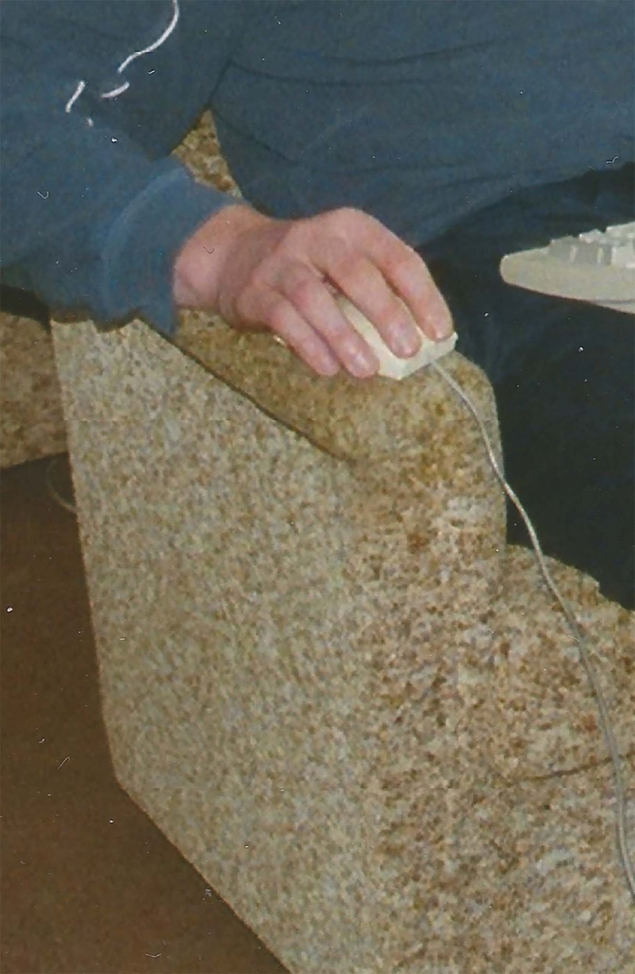 Fotografie eines Couchsessel auf dem ein Mensch sitzt mit einer weißen Computermaus in der Hand, der Bildausschnitt ist zur Hälfte abgeschnitten, sodass man nur einen Arm sieht