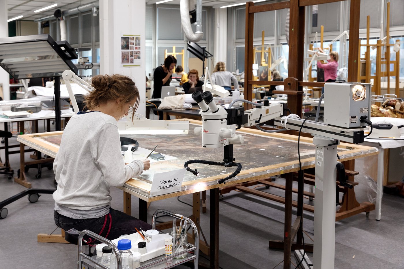 Links im Bild sitzt eine Studentin, die an einem Gemälde arbeitet; vor ihr liegt ein Gemälde auf dem Tisch, daneben steht ein großes Mirkoskop. Im Hintergrund sieht man andere Studentinnen, die an Gemälden arbeiten, teilweis an Staffeleien; dahinter die Fensterfront.