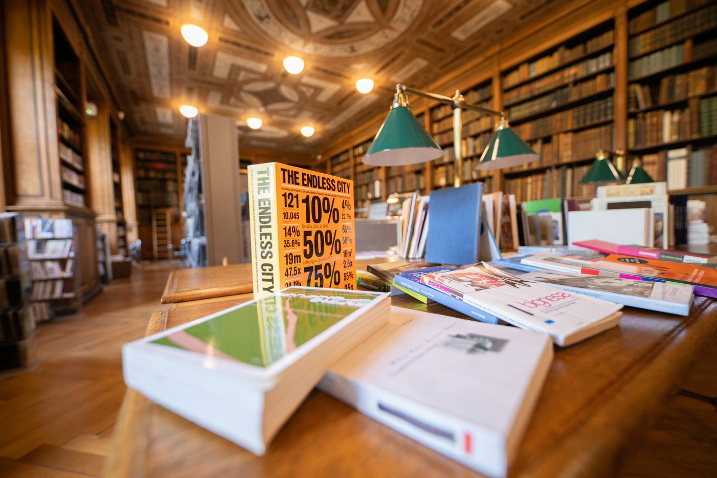 Die Farbfotografie im Querformat zeigt den von Theophil Hansen konzipierten Lesesaal mit historischem Buchbestand. Auf einem Originaltisch mit Leselampen im Vordergrund ist ein buntes Angebot von teils liegenden, teils stehenden Büchern präsentiert.