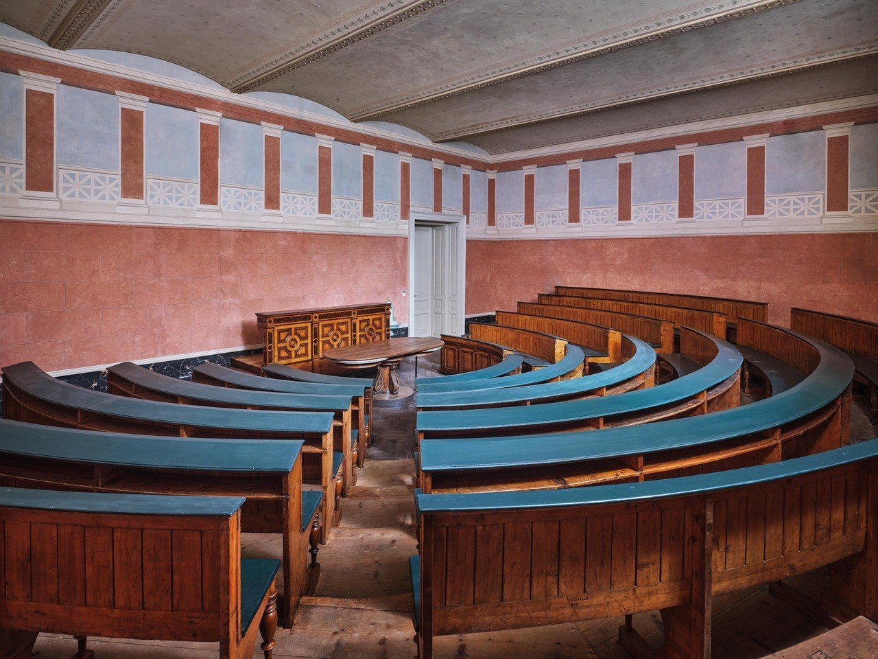 Fotografie eines Vortragssaales in dem die hölzernen Sitzbankreihen stufenförmig in einem Halbkreis angelegt wurden, in der Mitte ein Tisch und ein Rednerpult, die Wände sind in Terracotafarben mit illusionistischen Säulen bemalt, die von einem weißen Geländer unterbrochen werden.