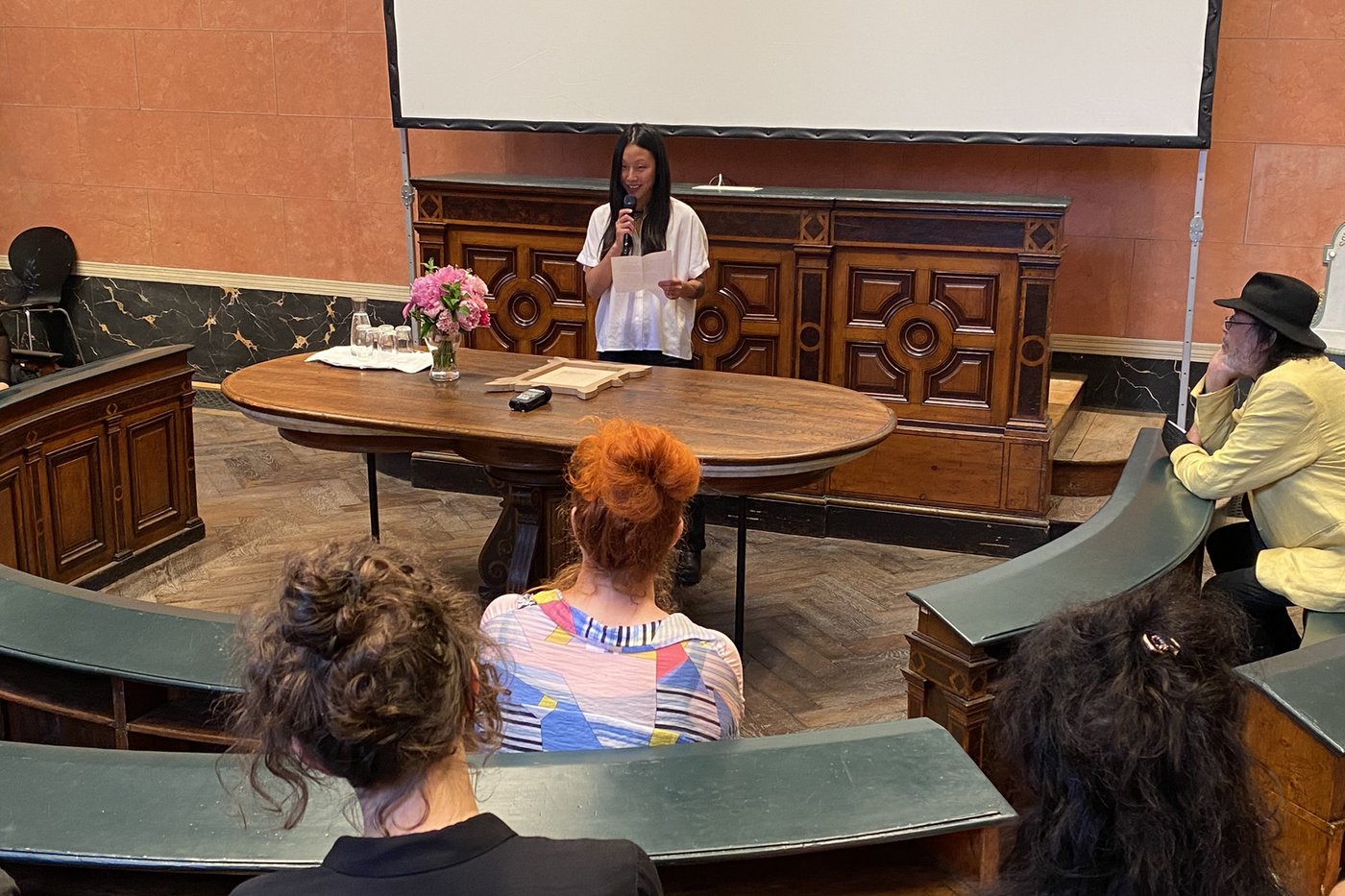 The prize winner Kyungrim Lim Jang stands at the speaker's table in the Anatomy Hall of the Academy of Fine Arts Vienna with a microphone in her hand and reads out her performative speech. On the table in front of her is her artistic work and a bouquet of flowers. Four people can be seen sitting backwards at the benches listening to her.