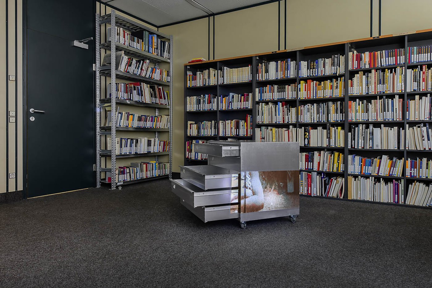 View of the exhibition in the archive room. In the background, bookshelves. In front of it, a mobile, low silver metal box with drawers pulled out. A film is projected on the side. On the still you can see a person with arms crossed in front of the chest.