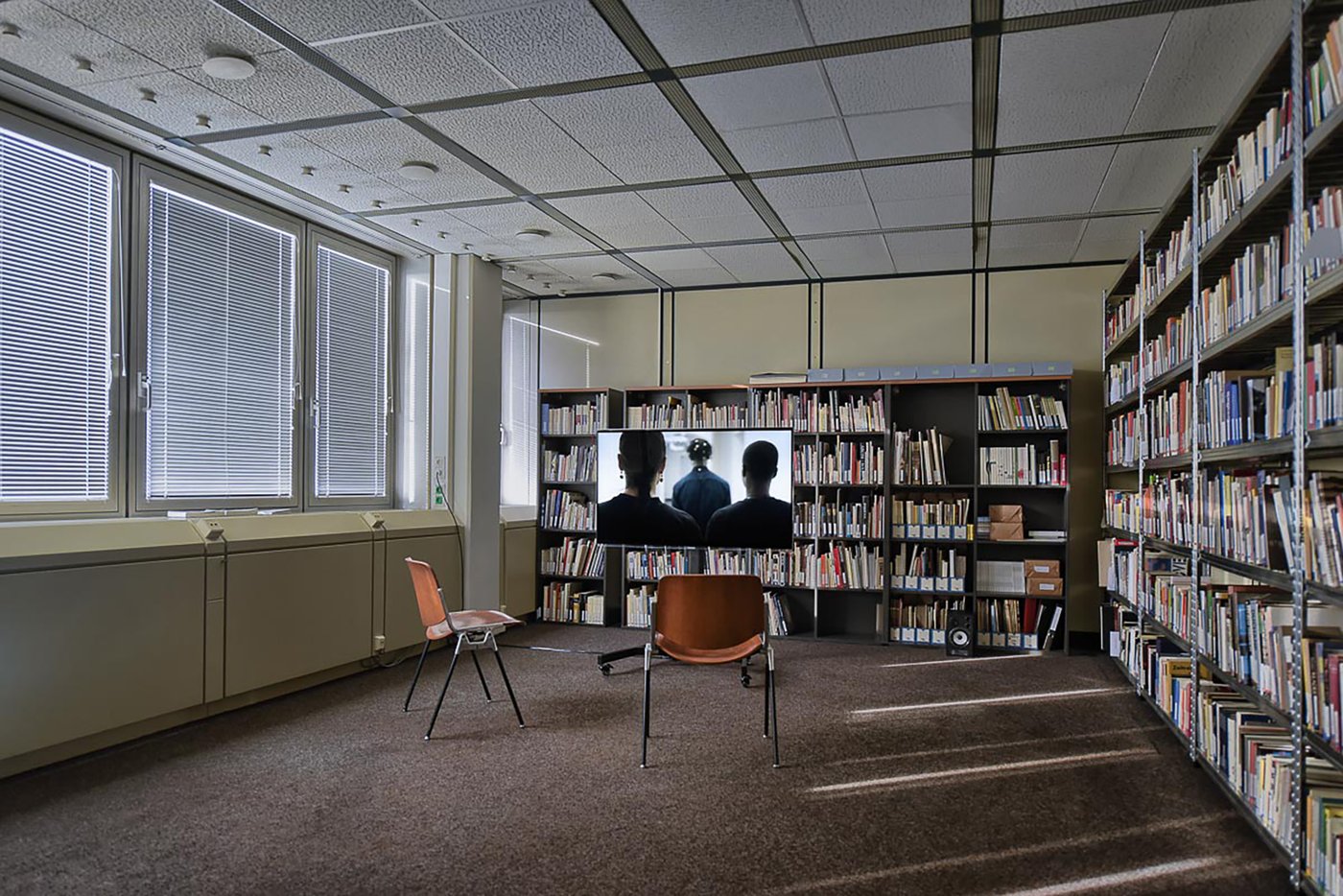 View of the exhibition in the archive room. In the background, bookshelves. In front of it, a mobile, low silver metal box with drawers pulled out. A film is projected on the side. On the still you can see a person with arms crossed in front of the chest.