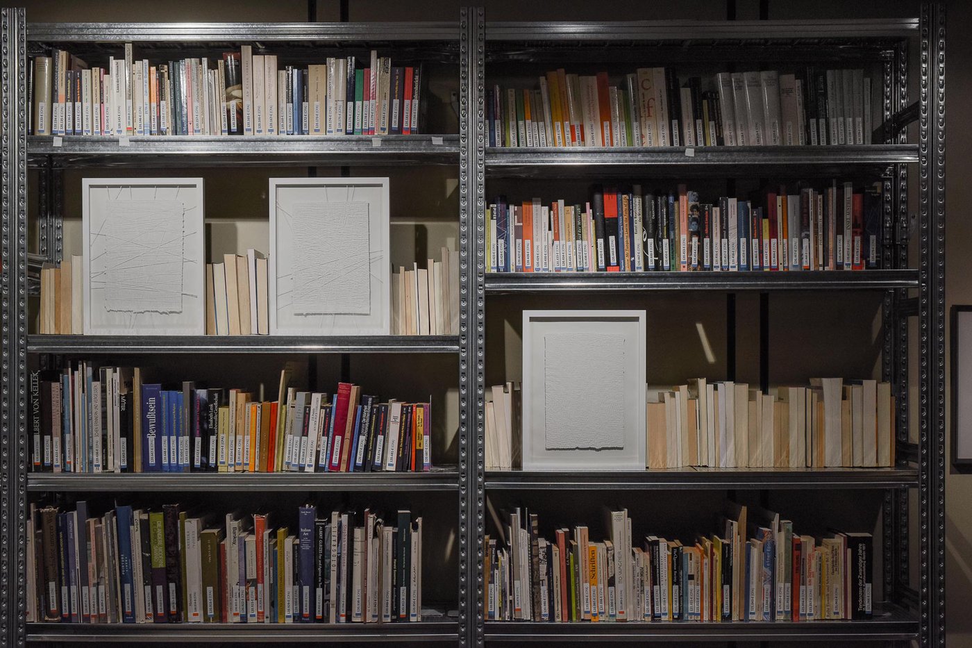 Metal bookcase with three framed white paper works presented in front of the books on the shelves.