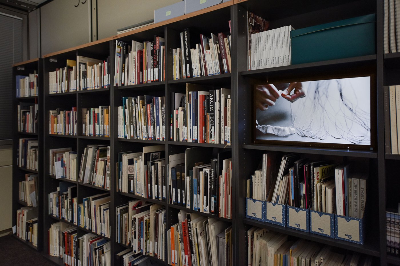 Bookshelf photographed at an angle, in one of the shelves a monitor can be seen running a video. On it, hands can be seen tying a paper thread.