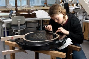 In the center of the image is a round, black lacquer tabletop with inlays, resting on two wooden work trestles. A student is sitting behind it on the right, bending over the object and working on the inlays.