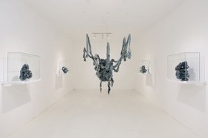 Exhibition view with blue-grey fabric bundles tied together to a large object and hanging from the ceiling, around small sotff bundles in glass showcases