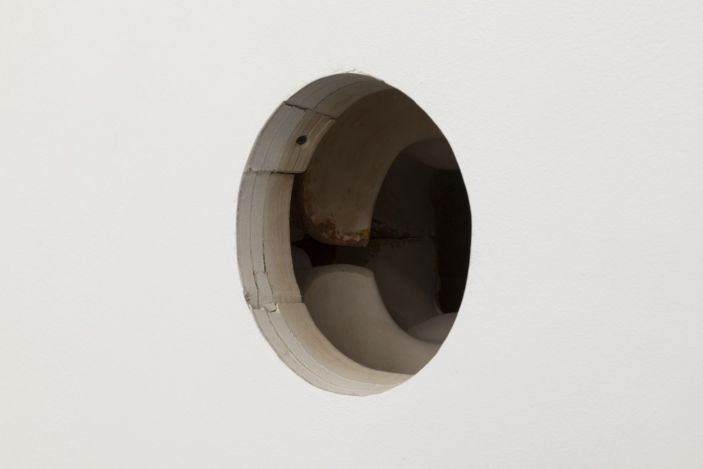 A hole in a white-painted plasterboard wall with two layers of plasterboard on a wooden structure underneath, in front of other objects that are difficult to capture.