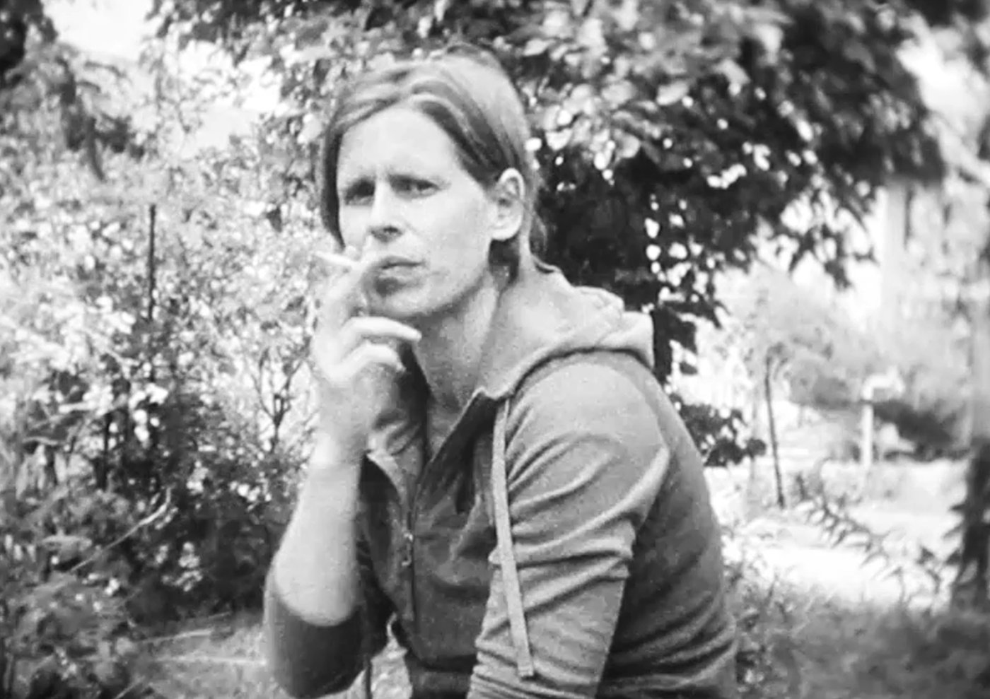 Black and white image of younger woman smoking cigarette with serious skeptical look. She is sitting in a garden.