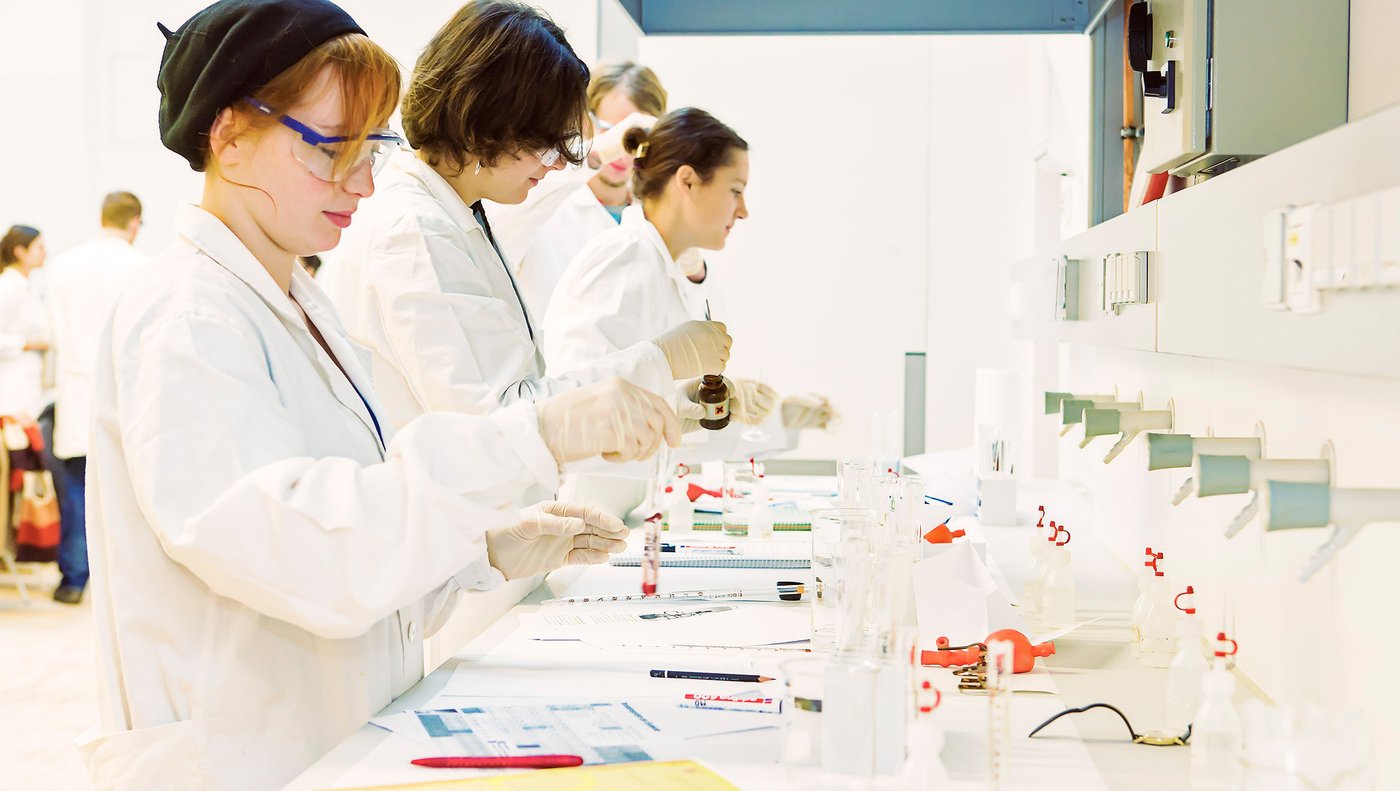 Students in white lab coats are standing at a lab bench. One person is holding a sample tube, another a small, brown bottle.