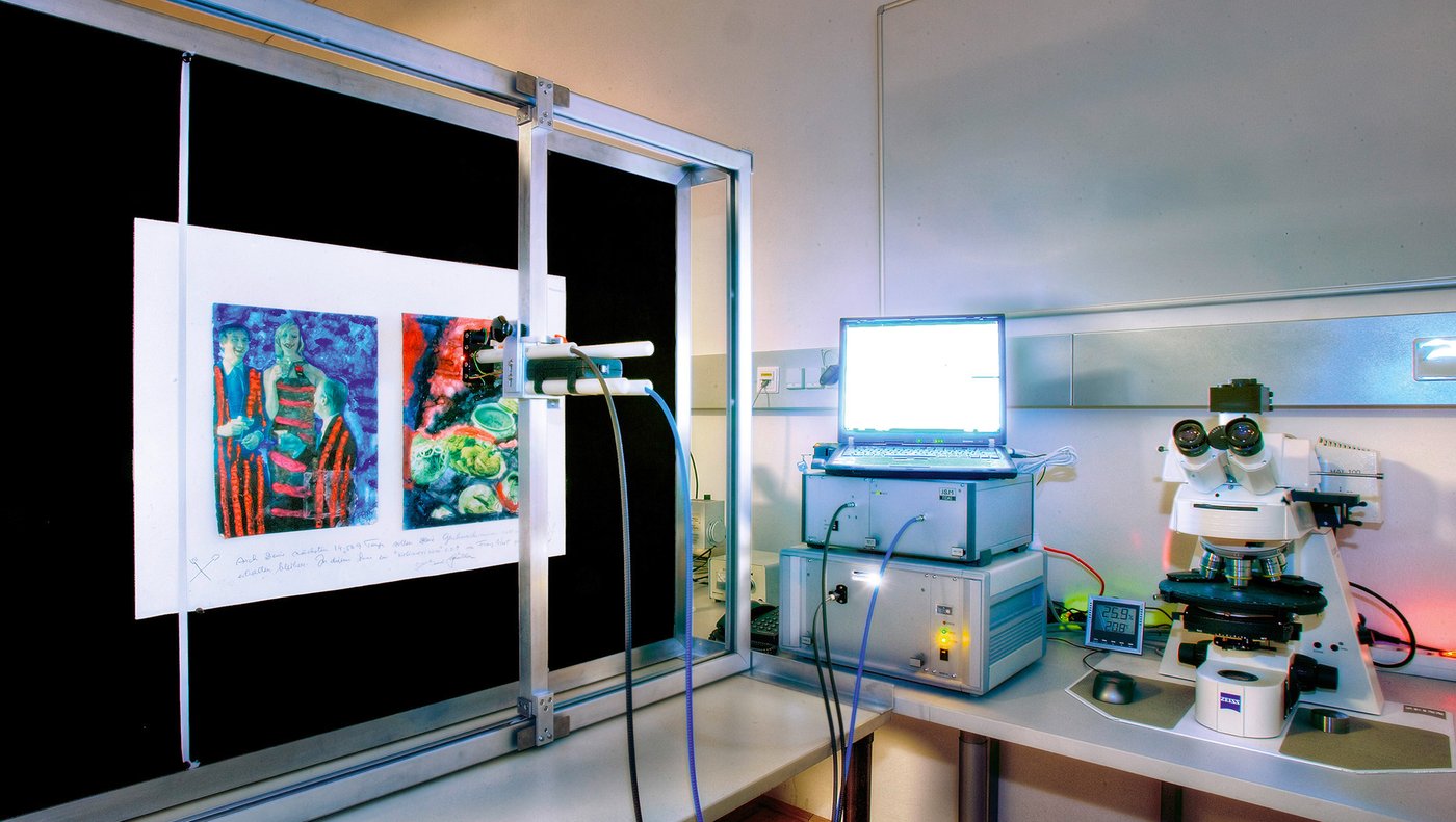 Two colourful pictures on a black background, behind metal scaffolding to which is mounted a camera-like device connected to a box and a laptop via cables. A microscope is standing next to this.