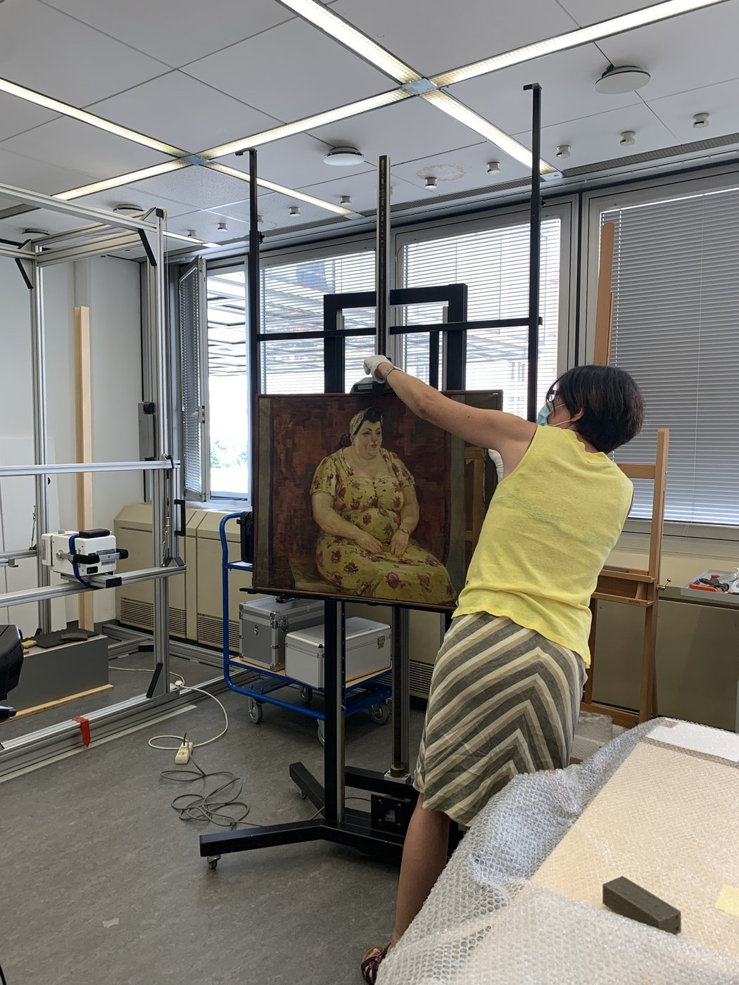 A person adjusting the height of picture on a wooden easel. The painting shows a curvy woman in a yellow dress with a pattern of red flowers and a matching headscarf on a brownish red background.