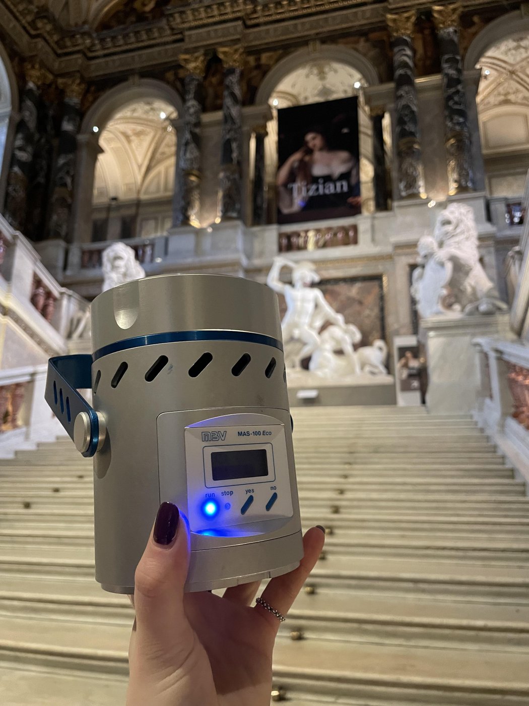 A hand holding a silver, cylindric container, in the blurry background there are stone steps, marble statues and a balustrade.