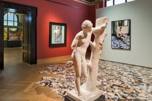 Exhibition view of an antique sculpture leaning on a piece of wood, in the background are Pictures on the wall and on the Floor, on the left side there is an open door into another room where you can see a the altar of Hieronimus Bosch