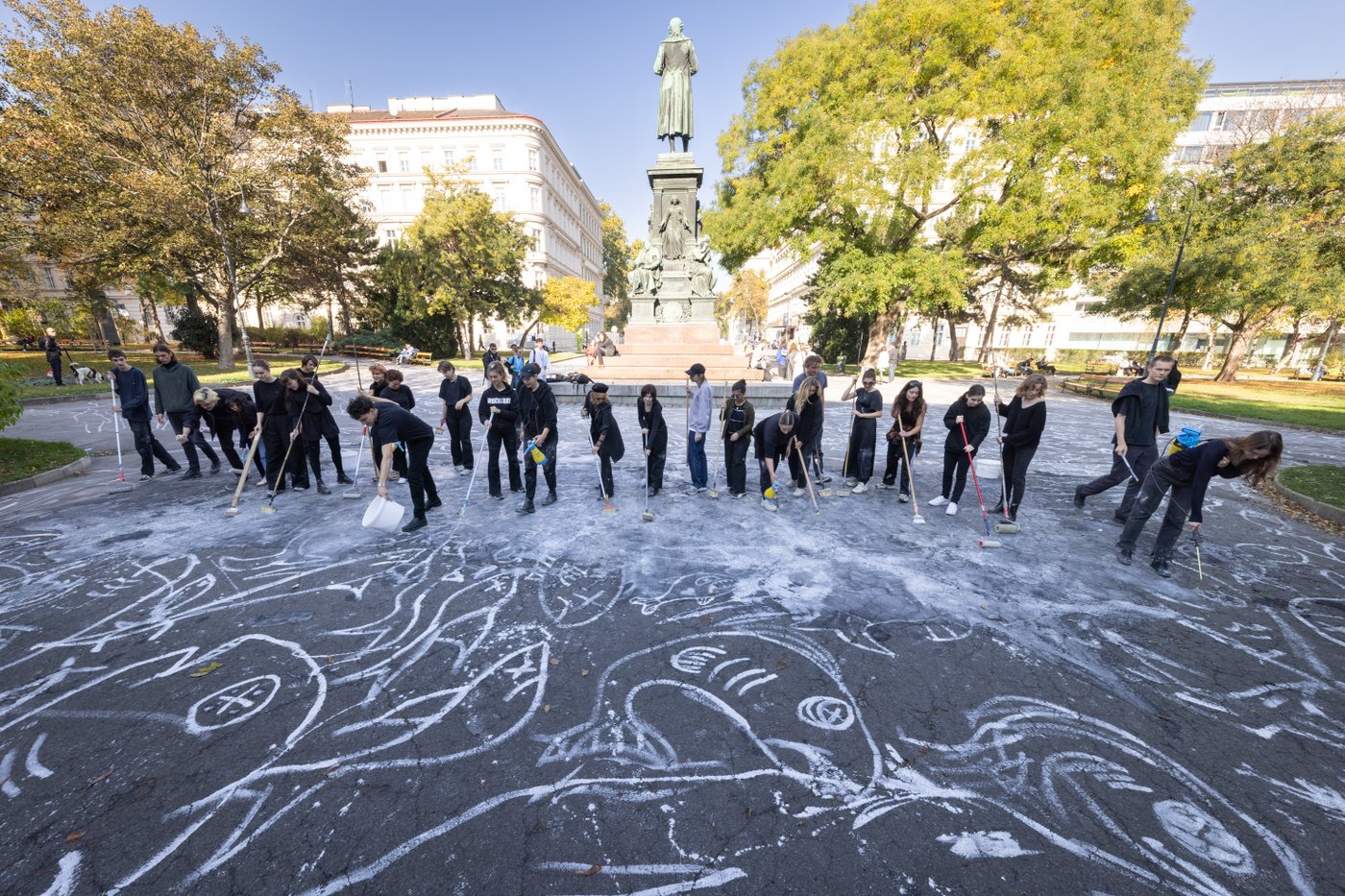 Several students working together to draw a chalk drawing with white paint and brooms on Schillerplatz.