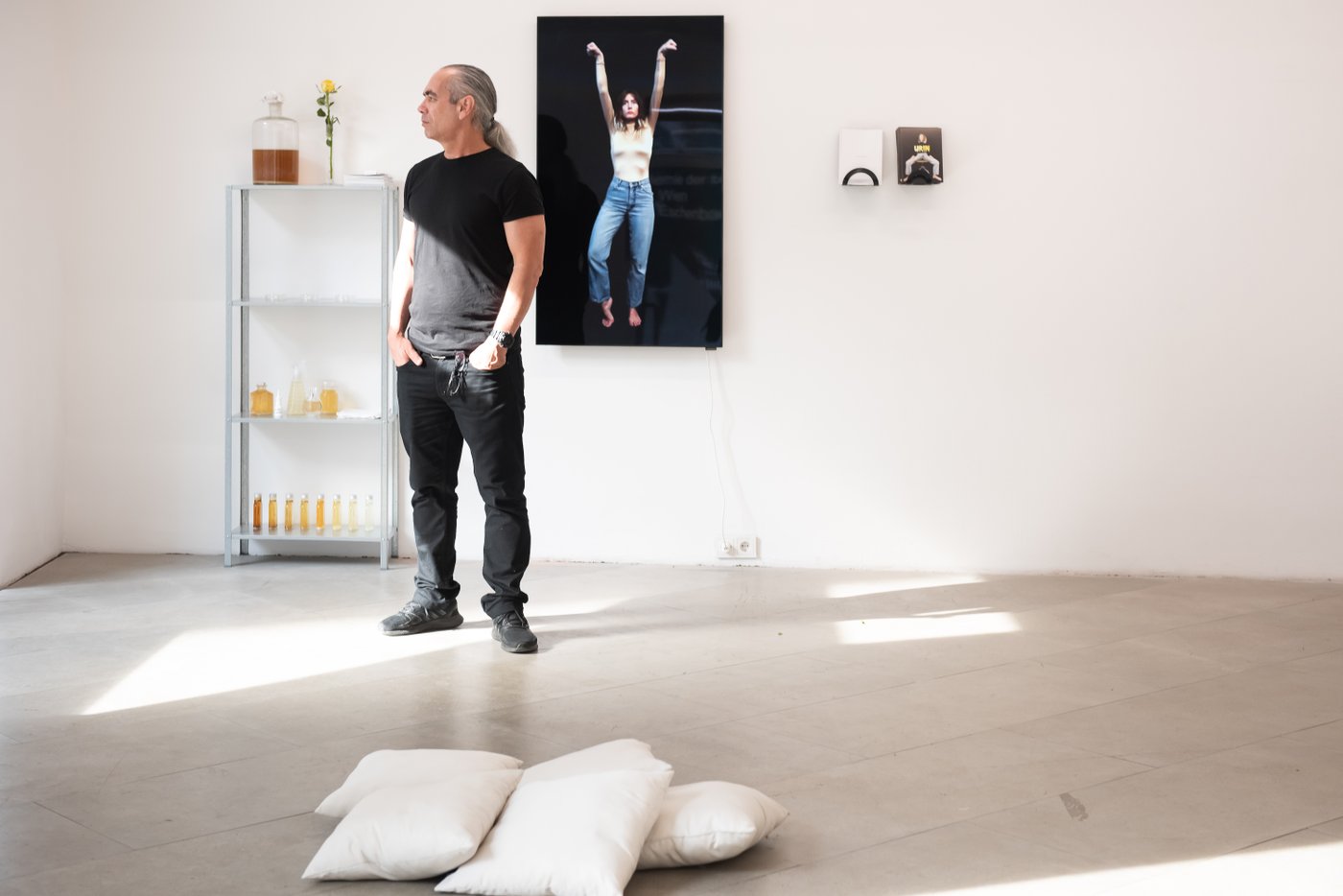 Person standing in the exhibition and looking outside the window, behind him bottles with a yellow liquid and a photo of a woman