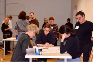 Several people are grouped around a table and are designing a poster in a workshop. In the background, other people are engaged in conversation.