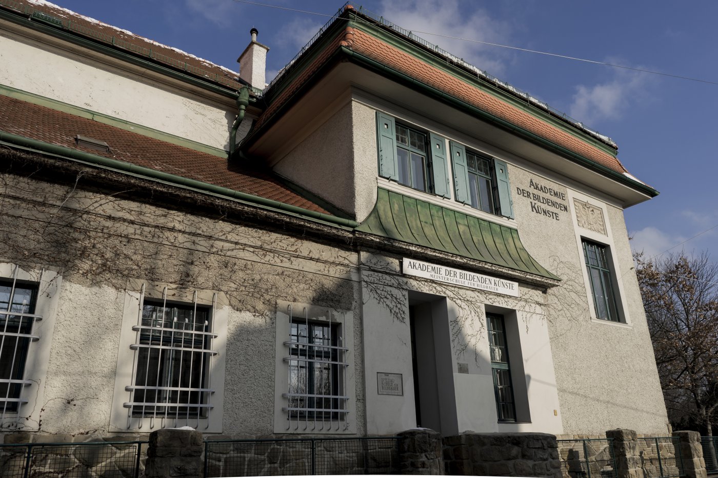 You can see the front of the building with various old and newer inscriptions, e.g. "Academy of Fine Arts Master School of Sculpture", "Special School of Sculpture".
Three windows have a grille, the portal has a green canopy and green shutters.