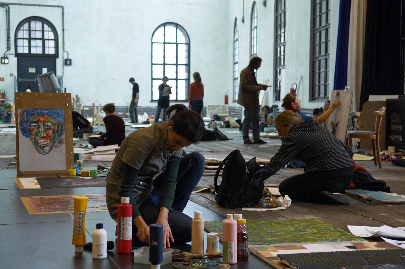 In the foreground a young woman sits on the floor, around her paint tubes, brushes and palette. She is absorbed in the work. Behind her on a wooden panel a hab finished picture of a portrait. In the background you can see many others in a similar situation standing or sitting at work.