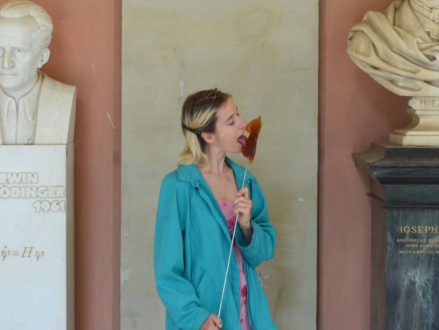 The artist stands between the busts of Erwin Schrödinger and Joseph Hyrtl in the arcade courtyard of the University of Vienna. She is holding a self-made lollipop in her hand, which is a 1:1 imprint of the mouth-nose area of Erwin Schrödinger's bust. The artist licks the lollipop with relish.