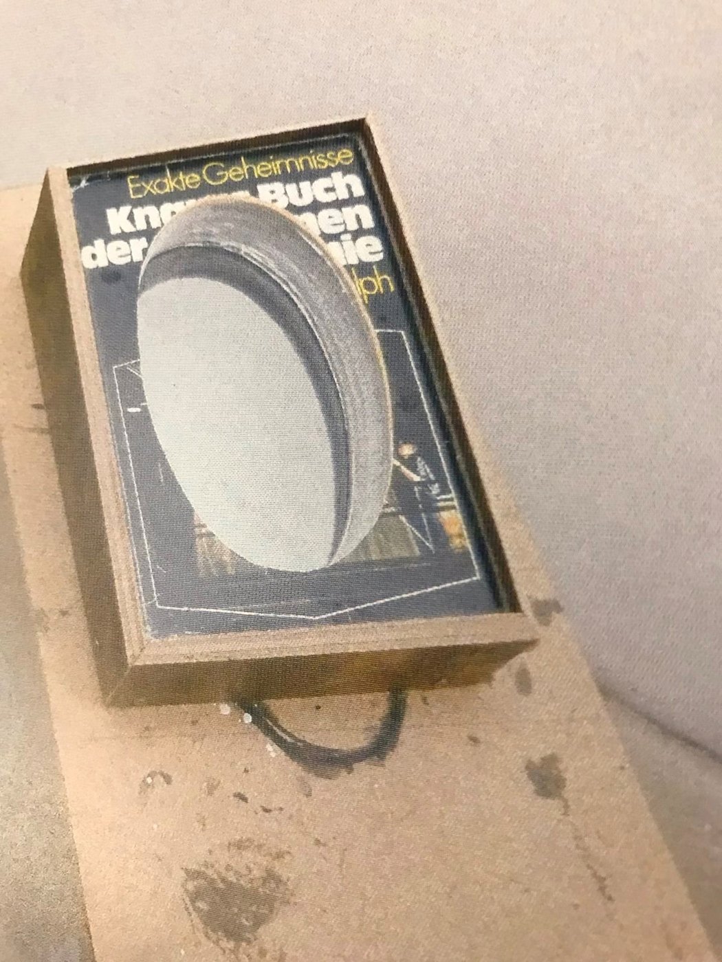 The color photograph shows a book in a rough frame attached to a larger wooden structure. A large, oval hole has been milled into the book so that the title can only be recognized in fragments: "Exakte Geheimnisse. Kn... Book of ... ie"