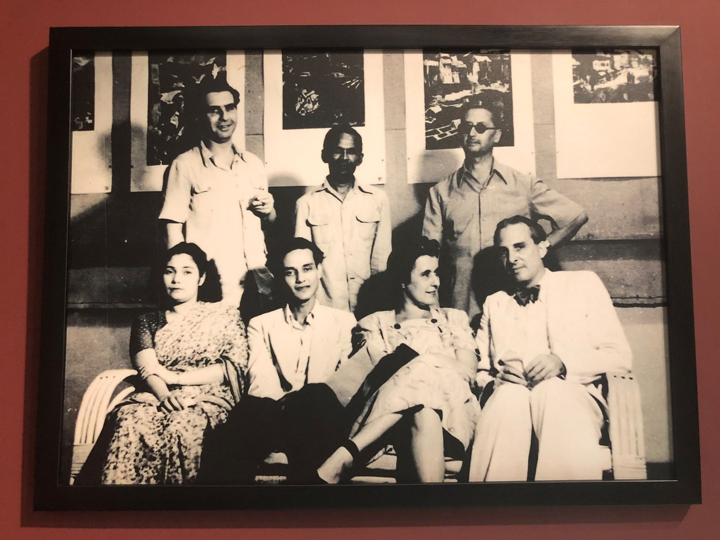 The black-and-white photograph shows a group of artists lined up in front of a row of graphic-art works. Sitting on a rattan bench in the foreground: unknown woman in a sari, Sayed Haider Raza, and Käthe Langhammer with her husband Rudi von Leyden. Standing behind them Walter Langhammer, K. H. Ara, and Emanuel Schlesinger (wearing sunglasses).